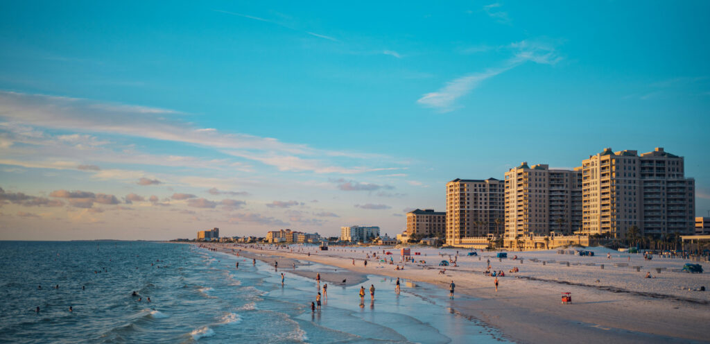 Beachfront with people walking, swimming, and buildings in the background under a blue sky.