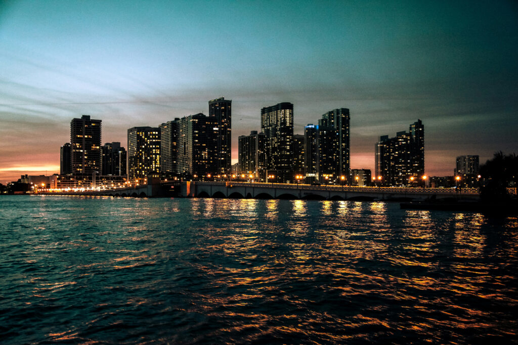 City skyline at dusk with illuminated buildings and bridge reflecting in the water.
