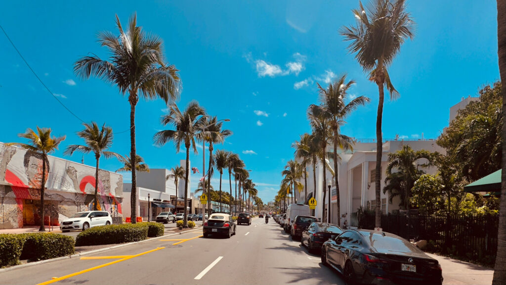 Street with palm trees, cars lining both sides, blue sky, mural on white building.