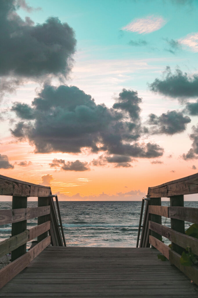 Wooden boardwalk leading to the ocean with a vibrant sunset and clouds in the sky.