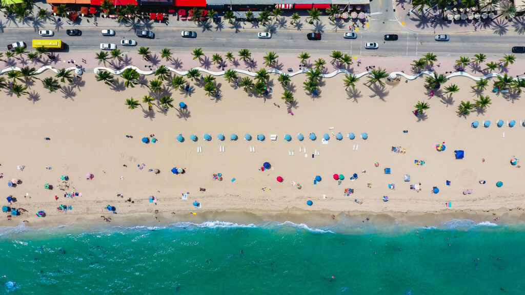 lance-asper-eJx43ng-fTU-unsplash Aerial view of a beach with colorful umbrellas, palm trees, and people by the ocean.