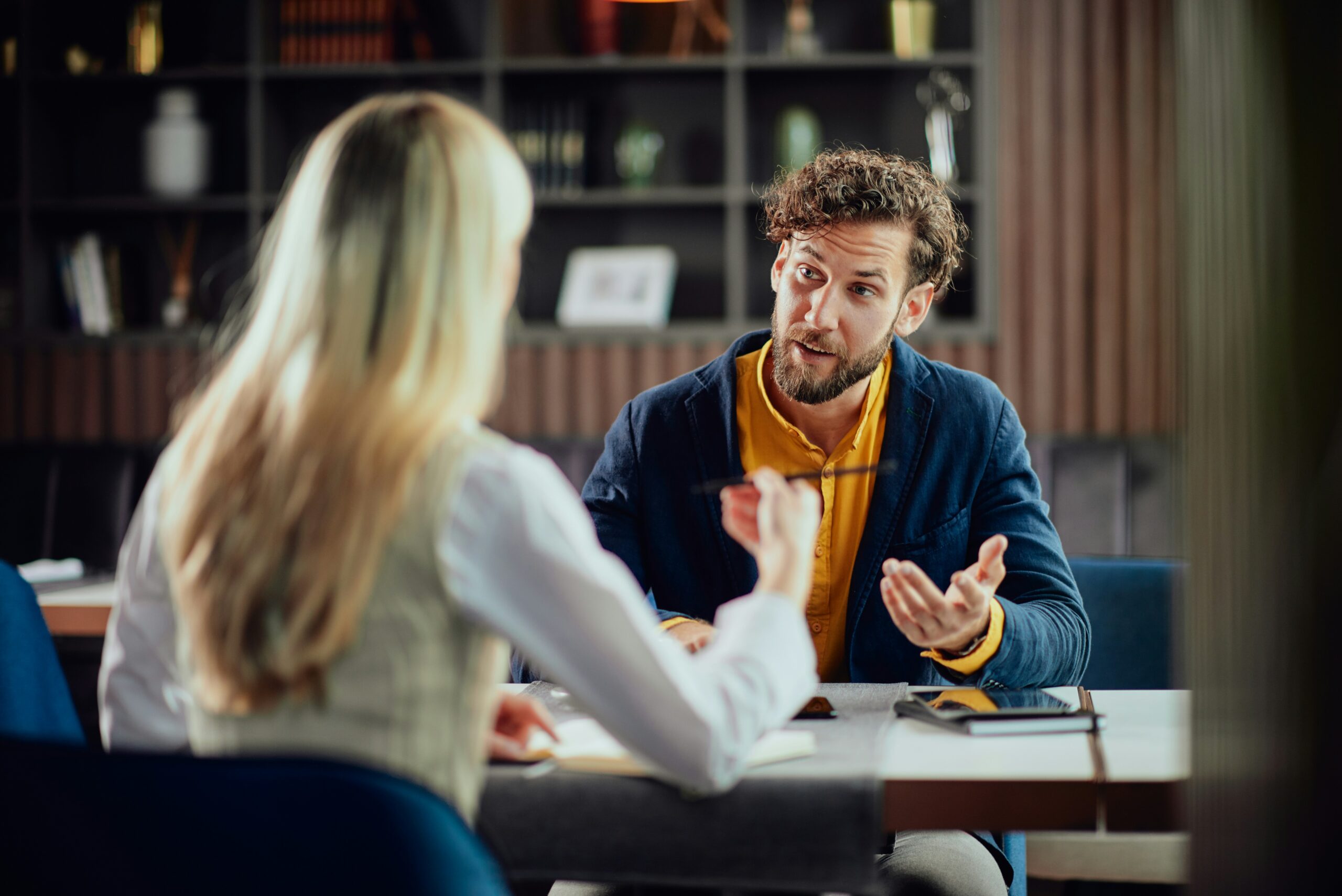Person in blue blazer and yellow shirt gesturing while talking to another person at a table.