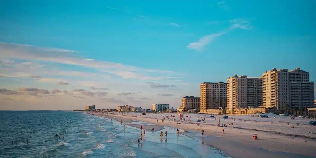 Beach scene with people along shoreline, large buildings in background, and blue sky above.