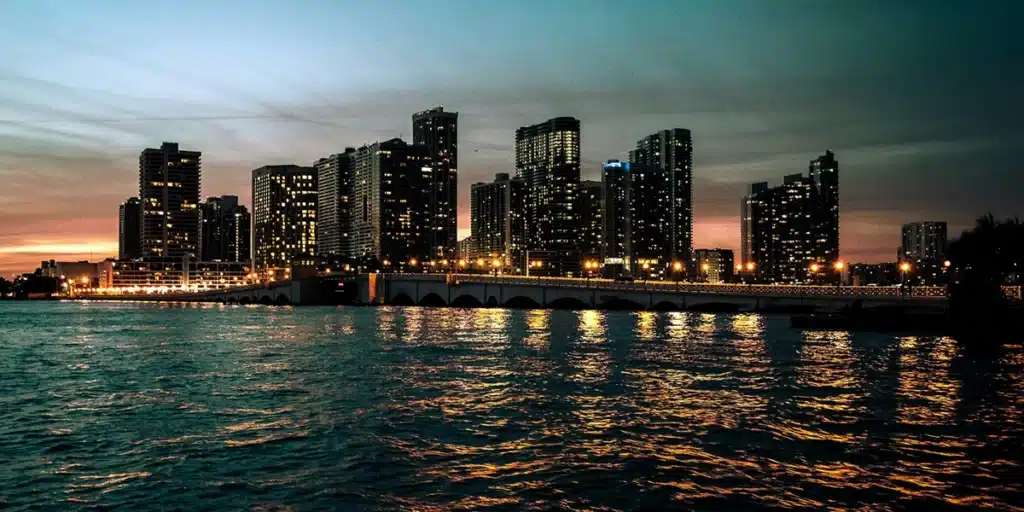 City skyline with illuminated buildings during dusk, reflecting on the water.