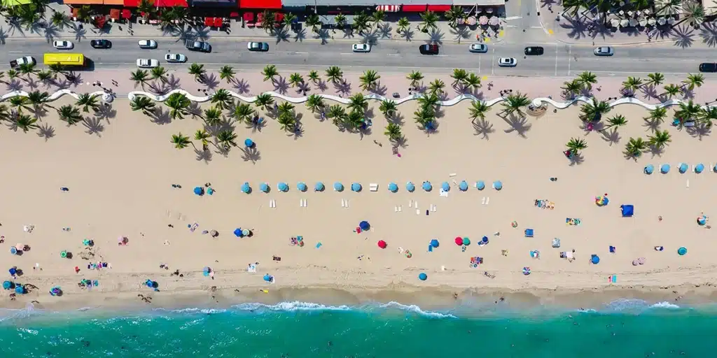 Aerial view of sandy beach with blue umbrellas, palm trees, and ocean waves, cars parked on road in background.